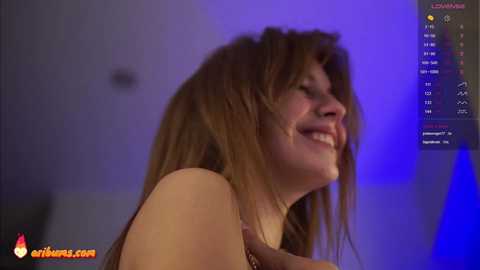 Media: Video of a smiling woman with long, wavy brown hair, under blue lighting, wearing a sleeveless top, captured in a candid moment.