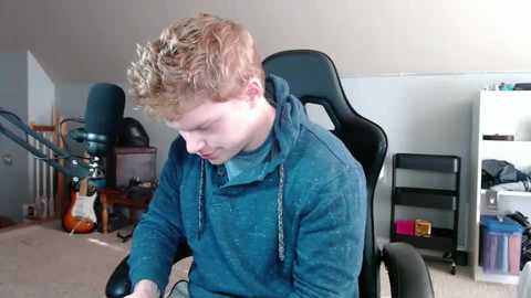 Media: Video of a young man with curly red hair, wearing a blue hoodie, seated in a black gaming chair, surrounded by a cluttered room with a guitar, speakers, and storage units.