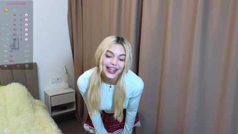 Media: Video of a smiling, blonde, young woman with long hair in a white top and red plaid skirt, bending slightly forward. Background includes a bed with a yellow blanket, beige curtains, and a calendar on the wall.