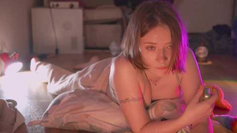 Media: Video of a young Caucasian woman with light skin, straight brown hair, and a small tattoo on her arm, lying on a bed in a dimly lit room, illuminated by pink and purple lights.