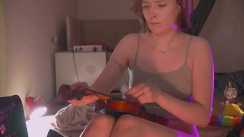 Media: Video of a woman with short hair, wearing a tank top and shorts, playing a violin in a dimly lit room with purple light, surrounded by cluttered furniture.