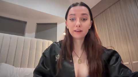 Media: Video of a light-skinned woman with long brown hair, wearing a black leather jacket, sitting on a white upholstered bed in a modern bedroom with beige walls and closed blinds.