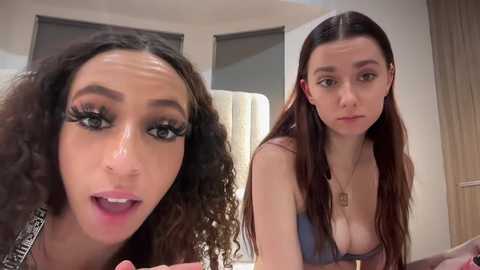 Media: Video of two young women with distinct appearances: curly-haired woman with dramatic makeup, and straight-haired woman with neutral expression, both indoors.