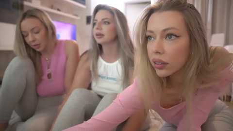 Media: Video of three young women in casual clothes, sitting indoors. Blonde-haired woman in pink sweater and light gray pants foreground, looking concerned. Two other women, one in white tank top, and another in light pink top, behind her.