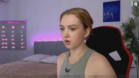 Media: Video of a young, fair-skinned woman with light brown hair in a ponytail, wearing a sleeveless green top, sitting in a red and black gaming chair. Background features a neatly made bed, a colorful LED strip, and a blue framed artwork.