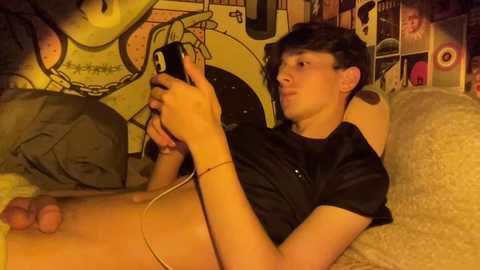 Media: Video of a young, slim, fair-skinned male with short dark hair, wearing a black shirt, playing with a phone, lying on a yellow couch in a cozy, dimly lit room.