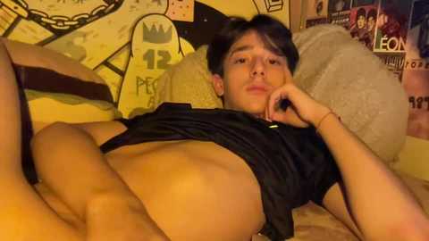 Media: Video of a young, slender, light-skinned man with short dark hair, lying on a bed with yellow walls adorned with posters. He is partially undressed, wearing only a black shirt, revealing his toned abdomen.