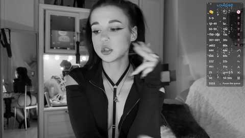 Media: Black-and-white video of a young woman with long hair in a ponytail, wearing a black jacket, with a serious expression, in a cluttered bedroom with a bed and a desk in the background.