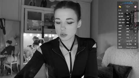 Media: A black-and-white video of a woman with light skin and dark hair in a ponytail, wearing a black jacket and makeup, leaning over a table in a dimly lit room. A computer screen shows a video call in the background.