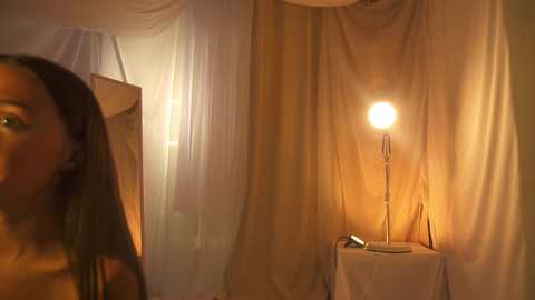 Media: A video of a woman with long, straight hair, partially visible, standing in a dimly lit room with soft, warm lighting, white draped curtains, and a small table with a lamp.