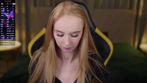 Media: A video of a young, fair-skinned, blonde woman with long hair, wearing a black shirt, sitting in a gaming chair. She appears focused, with a screen displaying a video game interface in the background.