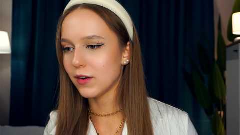 Media: Video of a young woman with fair skin, long brown hair, and a white headband. She wears a white jacket and gold necklace, with a dark green curtain and potted plant in the background.