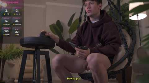 Media: Video of a young, pale-skinned man in a brown hoodie, sitting on a swing chair in a modern living room, holding a cup, surrounded by green plants, with a digital interface displaying health stats.