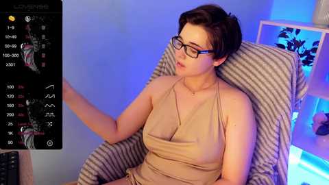 Media: Video of a woman with short brown hair, glasses, and beige halter top, seated in striped armchair, looking at phone with live streaming app interface, blue lighting.