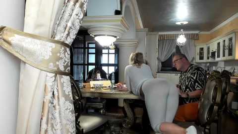 Media: A video depicts a grand dining room with a chandelier, ornate wallpaper, and elegant furniture. Two people, a woman in a white dress and a man in a black shirt, sit at a wooden table.