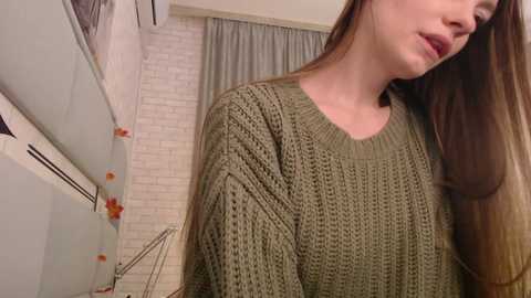 Media: Video of a young woman with long, straight brown hair, wearing a chunky, olive-green knitted sweater, standing indoors near a white brick wall and gray curtain.