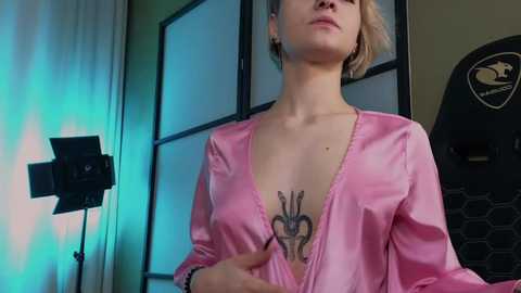 Media: Video of a fair-skinned woman with blonde hair, wearing a pink satin robe, revealing a heart tattoo on her chest. Background features a camera and studio lighting, suggesting a photoshoot setting.