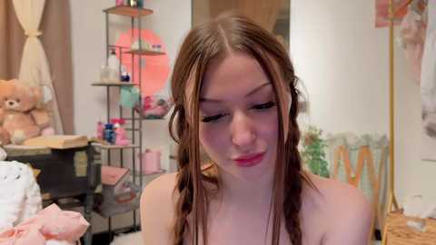 Media: A video of a young woman with long brown hair in braids, wearing pink lipstick, sitting in a cozy, cluttered bedroom with stuffed animals, shelves, and a mirror.