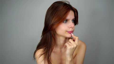 Media: Video of a young Caucasian woman with long, straight brown hair, light skin, and green eyes, looking contemplative while holding a pink lipstick to her lips. She's topless, with a plain, light grey background.