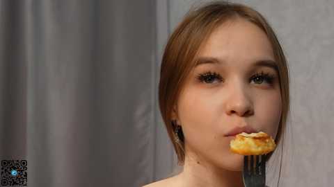 Media: A video of a young woman with light skin and straight brown hair, eating a sandwich with a fork in a neutral-toned room.