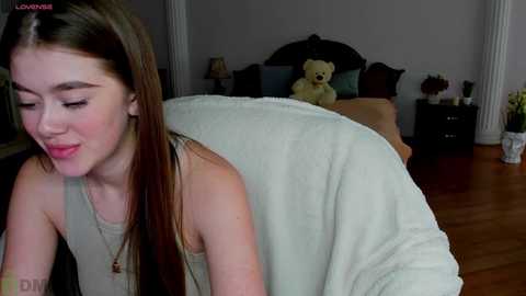 Media: A video of a young woman with long brown hair, wearing a light grey top, smiling while sitting on a bed with a beige blanket. The room has a wooden floor, a teddy bear, and dark furniture.