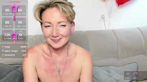 Media: Video of a smiling, topless blonde woman with freckles and sunburn on her chest, sitting on a beige couch, in a modern living room.