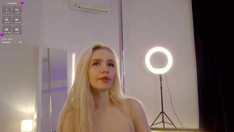 Media: A video of a topless, fair-skinned, blonde woman with long hair, in a dimly lit room with a glowing ring light.