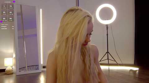 Media: Video of a nude blonde woman with long hair, sitting indoors. Background features a white wall, a large round light, and a mirror.