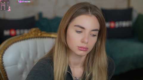 Media: Video of a young woman with long, straight blonde hair and fair skin, wearing a dark top, sitting on a green velvet sofa with a gold frame. Background features a blurred, ornate wallpaper and cushions.