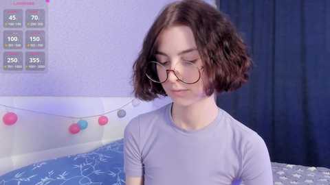 Media: Video of a young woman with short, curly brown hair, wearing glasses and a light purple t-shirt, in a pastel-colored bedroom with blue bedding, pink and red balloons, and a chart on the wall.