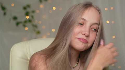 Media: A video of a young, fair-skinned woman with long, straight platinum blonde hair, wearing a light green necklace, sitting on a white chair, smiling while holding a lipstick. The background has soft, blurred bokeh lights and green ivy.