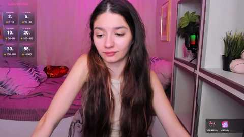 Media: Video of a young woman with long dark hair, light skin, and a slim figure, wearing a beige top, sitting in a pink-lit bedroom with a bed and green plants.