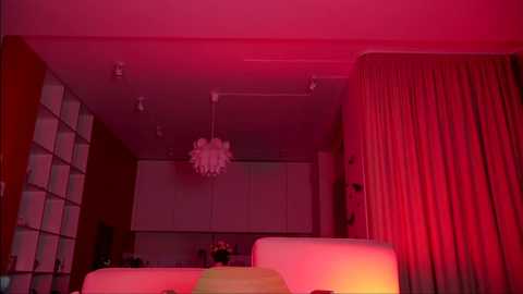 Media: Video of a modern, dimly-lit living room bathed in warm red lighting, featuring a white sectional sofa, a chandelier, and sheer curtains.