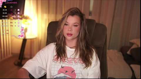 Media: Video of a young woman with long, wavy, light brown hair, wearing a white t-shirt featuring a pink shark graphic. She sits in a dark chair, with a glowing lamp and floral curtains in the background.