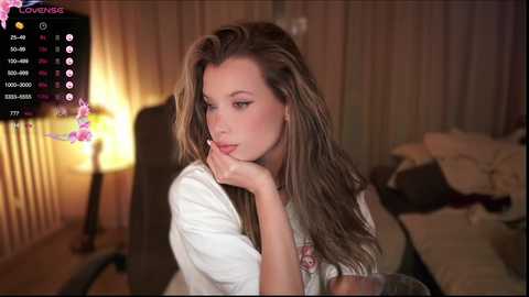 Media: Video of a young woman with long, wavy blonde hair, resting her chin on her hand, seated at a desk in a dimly lit room.