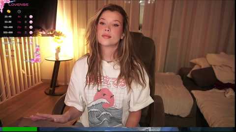 Media: Video of a young woman with long, wavy blonde hair, wearing a white graphic t-shirt, sitting in a dimly-lit room with a bed and a lamp.