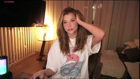 Media: Video of a young woman with long brown hair, wearing a white T-shirt with a pink flamingo design, sitting at a table in a dimly lit room with wooden walls, a TV, and a lamp.