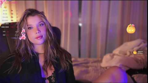 Media: Video of a young woman with light skin, long brown hair, wearing a black top and pink flower, sitting on a bed, surrounded by a Halloween-themed room with a pumpkin icon.