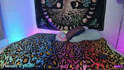 Media: Video of a cozy bedroom with a colorful, leopard-print duvet and a decorative tapestry featuring a tree and moon phases, lit by soft blue and purple LED lights.