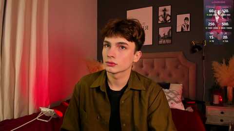 Media: Video of a young man with short dark hair, wearing a green shirt, sitting on a bed in a dimly lit room with red lighting, framed photos, and a TV screen displaying a game.