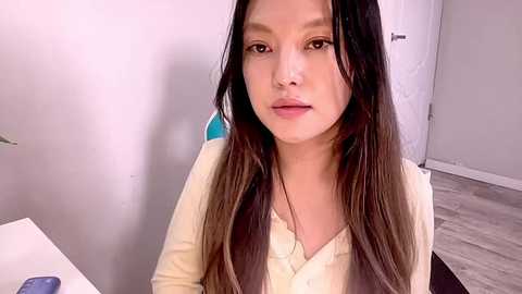 Media: Video of an East Asian woman with long black hair, fair skin, and a slight smile, wearing a beige cardigan, sitting indoors against a white wall and wooden floor.