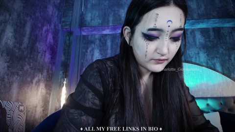 Media: Video of a young woman with dark hair and painted face, wearing a black leather jacket, in a dimly lit, industrial-style room with blue and purple tones.