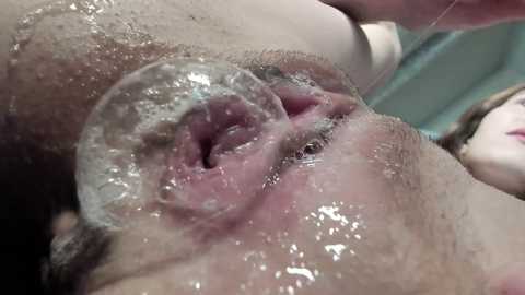 Media: A close-up video of a human vulva, partially submerged in water, with a clear, plastic-like substance covering part of the labia, revealing moist, pink inner tissues. The background shows a blurred, light-colored surface.