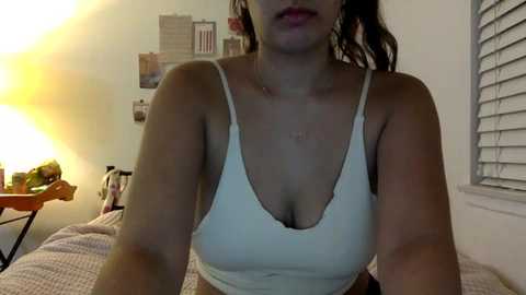 Media: Video of a light-skinned woman with long brown hair, wearing a white tank top, standing in a dimly lit bedroom with a bed, a lamp, and a window with blinds.