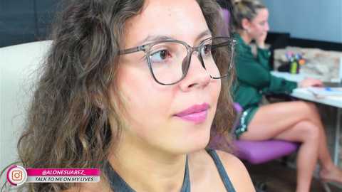 Media: Video of a young woman with curly hair, wearing glasses, pink lipstick, and a grey top, sitting in a modern, light-colored office.