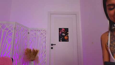 Media: Video of a woman with dark hair and visible tattoos, standing in a room with purple lighting. Behind her, a white door with a Halloween-themed sticker and a decorative screen.