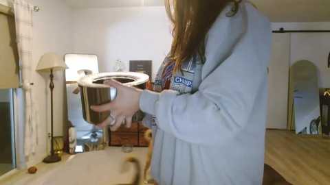 Media: Video of a woman in a light gray robe pouring water from a glass jug into a white sink. Background shows a bathroom with a mirror, lamp, and window.