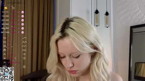 Media: Video of a blonde woman with fair skin and long, wavy hair, eyes closed, in a modern room with beige curtains, white walls, and a mirror.