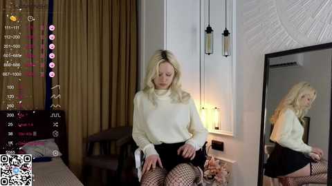 Media: Video of a blonde woman in a white sweater and black skirt sitting in a modern, dimly lit room with a large mirror reflecting her.