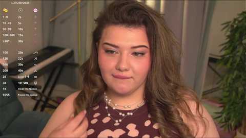 Media: A video of a plus-sized woman with wavy, shoulder-length brown hair and light skin, wearing a sleeveless brown top with a floral pattern. She's in a cozy, dimly lit room with a green plant in the background.